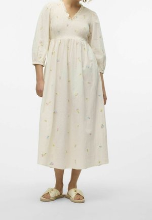 Vrouw draagt een crèmekleurige midi-jurk met driekwart mouwen en kleine pastelkleurige bloemenborduursels, gecombineerd met crèmekleurige slip-on sandalen.