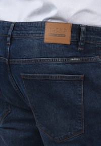 Vaqueros de mezclilla azul oscuro con una textura suave, que cuentan con un parche de cuero marrón etiquetado "BLEND" en la cintura y un bolsillo trasero estándar.
