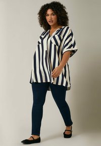 Túnica a rayas en crema y azul marino, con mangas cortas, cuello en V y dos bolsillos frontales, combinada con jeans oscuros ajustados y bailarinas.