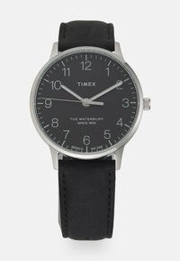 Zegarek Timex Waterbury z czarną tarczą, srebrną stalową kopertą i czarnym skórzanym paskiem. Cechy obejmują numeryczne znaczniki godzinowe oraz wyświetlacz daty.