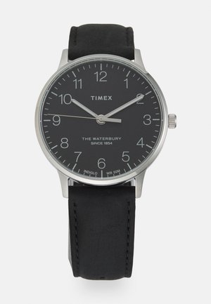 Hodinky Timex Waterbury s čiernym ciferníkom, strieborným puzdrom z nehrdzavejúcej ocele a čiernym koženým remienkom. Medzi funkcie patrí numerické hodinové marker a zobrazenie dátumu.