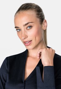 Marineblaues, knopfgeschlossenes Kleidungsstück mit hohem Kragen, langen Ärmeln und ausgeschnittenem Akzent an den Manschetten. Glatte Textur. Das Modell berührt sanft das Handgelenk.