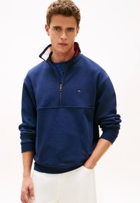 Μπλε ναυτικό πουλόβερ με γιακά μισής φερμουάρ, διαθέτει ριμπ μανσέτες και τελείωμα. Μικρό λογότυπο στο στήθος· το ύφασμα φαίνεται μαλακό με λεία υφή.