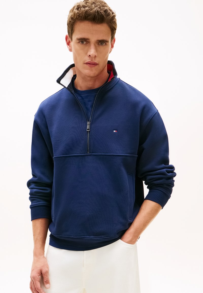 Μπλε ναυτικό πουλόβερ με γιακά μισής φερμουάρ, διαθέτει ριμπ μανσέτες και τελείωμα. Μικρό λογότυπο στο στήθος· το ύφασμα φαίνεται μαλακό με λεία υφή.