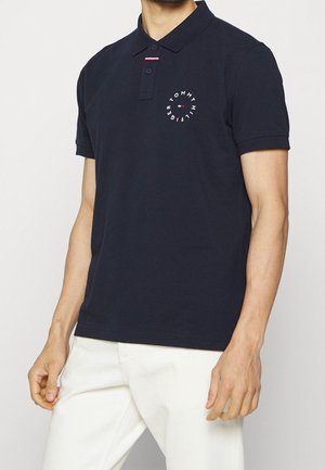 Mand iført en mørkeblå polo med et rundt Tommy Hilfiger-logo på venstre bryst og hvide bukser.