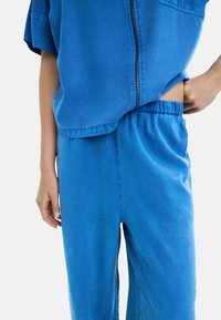 Osoba w niebieskiej bluzie na zamek i dopasowanych spodniach z elastycznym pasem, z jedną ręką opartą na biodrze, a drugą zwisającą kolo ciała.