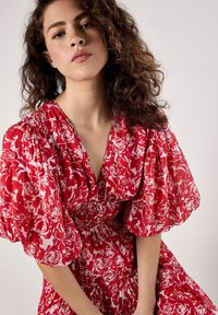 Rotes Blumenkleid mit Puffärmeln, tiefem V-Ausschnitt und gerafftem Bund. Der Stoff hat ein leichtes, strukturiertes Aussehen mit weißen Rosenmustern.