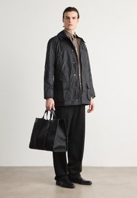 Veste légère noire avec fermeture éclair à l'avant, deux poches et col bleu marine. Associée avec un pantalon noir et un sac fourre-tout texturé noir.