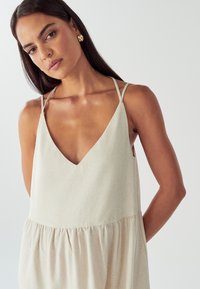 Moteris su ilgomis tamsiomis plaukais, vilkinti be rankovių smėlio spalvos suknelę su plonomis dvigubomis petnešomis ir auksiniais žiedais ausyse, prieš vienodą foną.