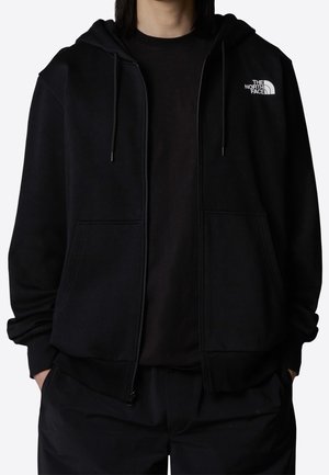 Musta vetoketjullinen huppari, joka on valmistettu pehmeästä kankaasta. Siinä on kiristysnauhallinen huppu, resorit hihansuissa ja pieni valkoinen logo vasemman rinnan yläpuolella.