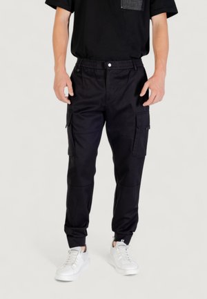 Zwarte cargobroek met elastische tailleband, meerdere zakken, taps toelopende pasvorm en een gladde stofstructuur. Gecombineerd met witte sneakers.