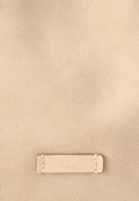 Μπεζ σουέντ ύφασμα με απαλή υφή, με ορθογώνια δερμάτινη ετικέτα με ανάγλυφα γράμματα "Gianni Chiarini Firenze" σε χρυσό χρώμα.