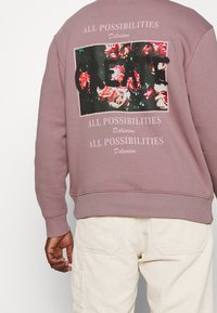 Sweatshirt rose poussiéreux avec un motif floral et du texte au dos. Poignets et ourlet côtelés. Fabriqué en tissu doux et texturé.