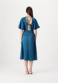 Robe teal avec des manches courtes à volants, dos nu avec des bretelles croisées, et une jupe évasée. Texture lisse, semblable à du satin. Talons légers.