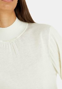 Crème kleurige gebreide trui met een hoge hals, voorzien van pintuck-details en kleine parelaccenten langs de kraag. Zachte textuur.