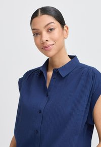 Camicia blu navy con maniche corte, chiusura a bottoni e design a righe testurizzate con colletto. Presenta bottoni frontali e tessuto morbido.