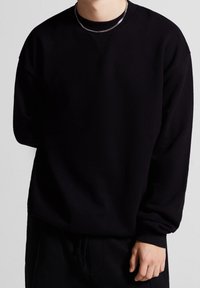Personne portant un sweat-shirt noir uni à manches longues et un pantalon noir, avec une chaîne en argent visible autour du cou.
