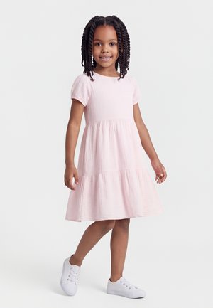 Mlada deklica stoji in se smehlja, oblečena v svetlo rožnato kratek rokav obleko in bele superge, na čistobelem ozadju.