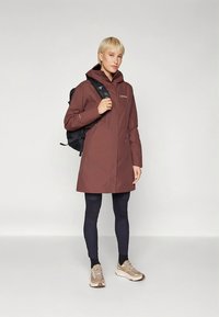 Vanntett brun parkasjakke med hette, modellert med svart ryggsekk, kombinert med svarte leggings og beige joggesko. Jevn tekstur, minimalistisk design.