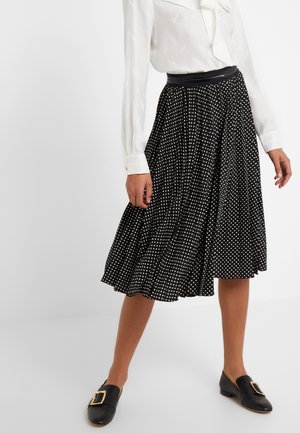 Femme portant une jupe midi plissée noire à pois blancs, un chemisier blanc à manches longues et des mocassins noirs avec des boucles dorées.