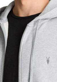 Felpa grigia con zip e cordino, caratterizzata da un discreto logo di teschio di capra sul petto. T-shirt nera visibile sotto. Tessuto morbido.