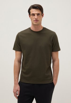 Hombre con cabello corto y oscuro que lleva una camiseta de manga corta verde oliva oscura y pantalones negros, sobre un fondo claro y liso.