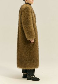 Kobieta w długim brązowym futrze, nosząca czarne spodnie i czarne mokasyny, stojąca bokiem na jednolitym jasnym tle.