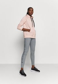 Felpa con cappuccio rosa chiaro con tasca frontale, abbinata a leggings grigi con branding laterale e scarpe da ginnastica nere con suole bianche.