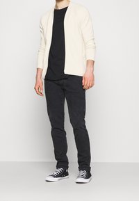 Mann in einem weißen offenen Cardigan, schwarzem T-Shirt, schwarzen Slim-Fit-Jeans und schwarzen High-Top-Sneakern, der vor einem schlichten Hintergrund steht.