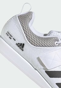 Zapatilla deportiva blanca con parte superior de malla texturizada, acentos grises y una suela de goma blanca. Presenta un logo en relieve y detalles de altura de apilamiento.