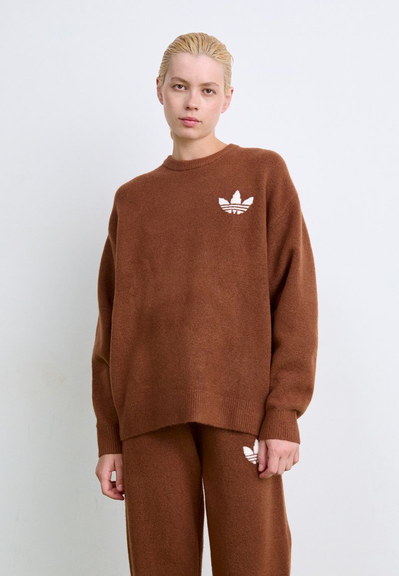 Brun strikket genser med avslappet passform, ribbestrikkede ermekanter og nederkant. Har en liten hvit logo på venstre bryst. Materialet virker mykt og teksturert.