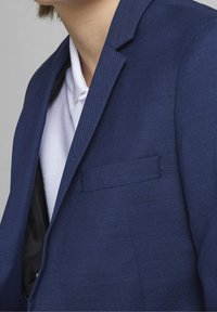 Granatowy żakiet z teksturowanej tkaniny, slim fit, z wywijanymi klapami i jedną kieszenią na piersi. Nosi się go na białej koszuli.