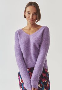 Violetter Strickpullover mit V-Ausschnitt und langen Ärmeln, der eine weiche Textur und ein geripptes Muster aufweist, kombiniert mit einem mehrfarbigen gemusterten Rock.