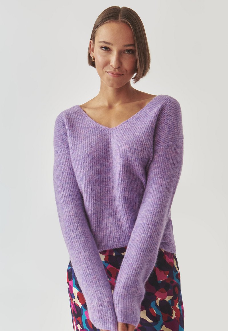 Violetter Strickpullover mit V-Ausschnitt und langen Ärmeln, der eine weiche Textur und ein geripptes Muster aufweist, kombiniert mit einem mehrfarbigen gemusterten Rock.