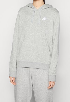 Osoba ubrana w jasnoszarą bluzę z kapturem Nike i pasujące spodnie dresowe, stojąca na tle jednolitego jasnego tła.