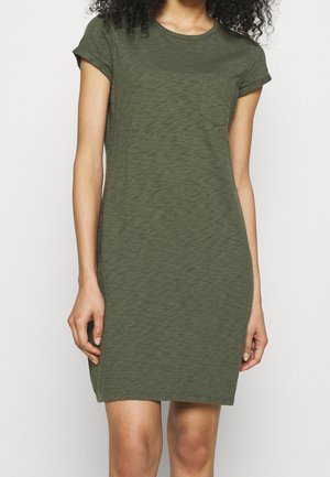 Femme portant une robe t-shirt manches courtes de couleur vert olive avec une petite poche poitrine, debout devant un fond clair uni.