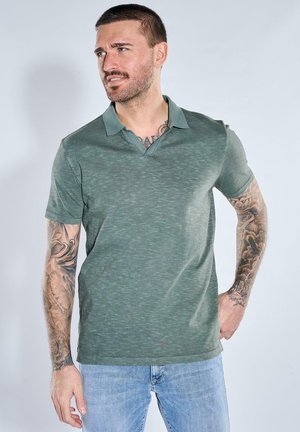 Mann mit kurzen Haaren und Tätowierungen an beiden Armen trägt ein grünes kurzärmeliges Poloshirt und hellblaue Jeans, schaut zur Seite.