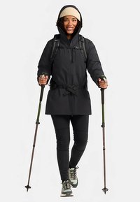 Giacca impermeabile nera con chiusura a zip e fibbia, indossata sopra leggings neri e berretto beige. Bastoncini da trekking e stivali robusti completano l'outfit.