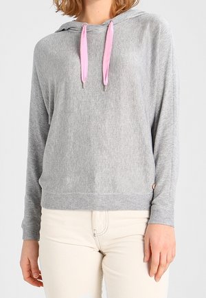 Femme portant un sweat à capuche gris clair avec des cordons roses et un pantalon écru, debout devant un fond blanc uni.