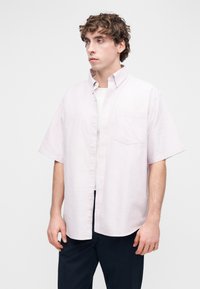 Νέος άνδρας με σγουρά μαλλιά που φοράει ανοιχτό ροζ πουκάμισο με κοντά μανίκια πάνω από λευκό μπλουζάκι και σκούρο παντελόνι, κοιτάζοντας προς τα αριστερά.