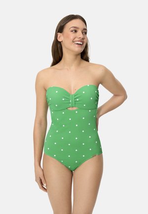 Mujer sonriendo, vestida con un bañador de una pieza sin tirantes verde con lunares blancos y un pequeño corte delantero, de pie frente a un fondo liso.