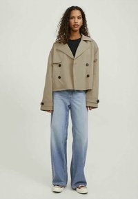 Casaco cropped bege de corte duplo com grandes botões pretos, gola larga e descontraída, combinado com calças de ganga de perna larga de tom azul claro.