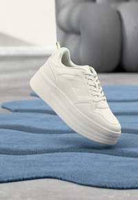 Balti platforminiai sportbačiai iš tekstūruotos odos, su storu padu, apvalia nosimi ir iškilimais šonuose, sklendžiantys virš mėlynos kilimėlio.