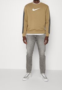 Beige sweatshirt med vitt Nike-logotyp, har svartvita randiga detaljer på ärmarna. Bärs med ljusgrå jeans och grå sneakers.