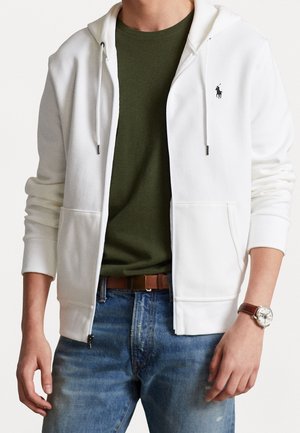 Hombre con sudadera blanca con cremallera y logo negro, camiseta verde oliva, vaqueros azules, cinturón marrón y reloj de pulsera, de pie con las manos relajadas.