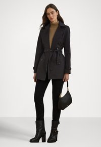 Czarny płaszcz typu trencz z paskiem, o podwójnym rzędzie guzików, z detalami guzików, noszony na brązowym golfie, w zestawie z czarnymi spodniami i botkami na obcasie.