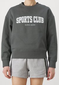 Felpa in cotone grigia con la stampa in bianco di "SPORTS CLUB" e "BJÖRN BORG". Presenta un colletto rotondo e una vestibilità comoda. Abbinata a pantaloni corti grigi.