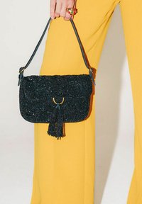 Borsa nera con perline, nappina e dettagli dorati, dotata di chiusura a patta e sottile tracolla, tenuta da una mano sopra pantaloni gialli.
