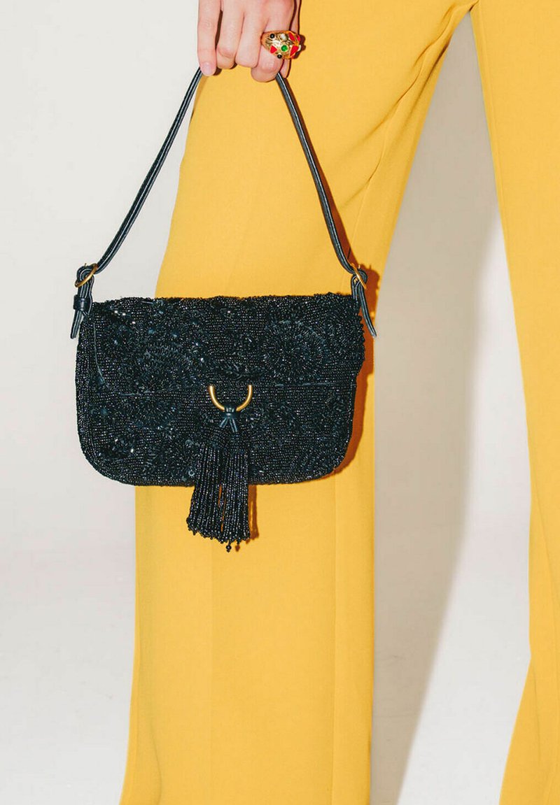Borsa nera con perline, nappina e dettagli dorati, dotata di chiusura a patta e sottile tracolla, tenuta da una mano sopra pantaloni gialli.