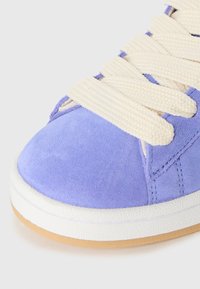Lavender suede sneaker features creamy flat laces, white rubber midsole, and light brown outsole with textured detail.